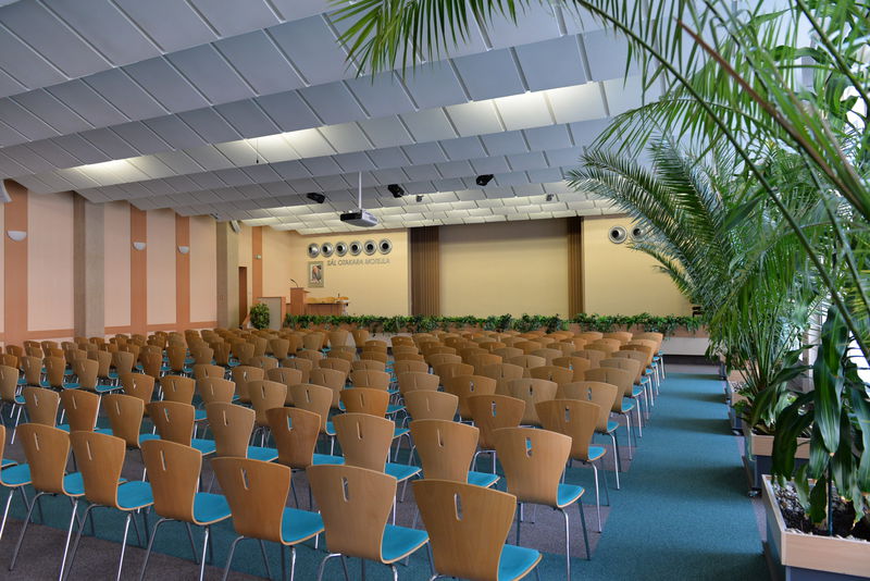 Na fotografii je zobrazen velký sál. Jsou v něm rozestavěny židle v řadách a na straně místnosti jsou pokojové rostliny.